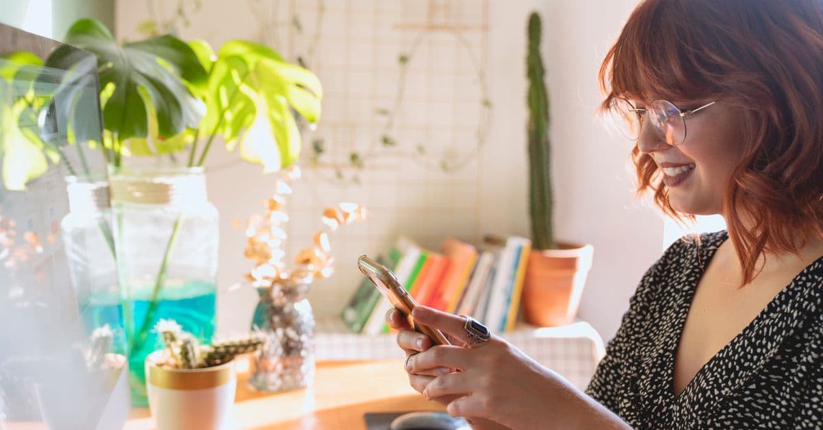 A woman with red hair smiles while using a smartphone in a bright and cozy home office.