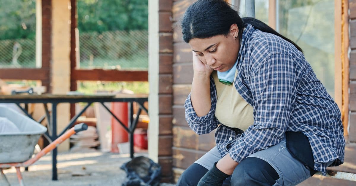 An African American woman in a plaid shirt sits, appearing tired at a construction site.
