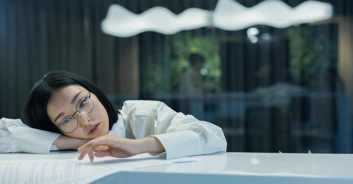 Asian woman in glasses rests her head on a counter with papers, showing signs of fatigue.