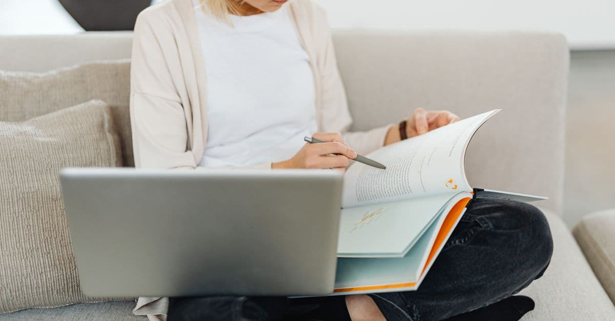 Woman sitting cross-legged on sofa, studying with a textbook and laptop, creating a cozy learning environment.