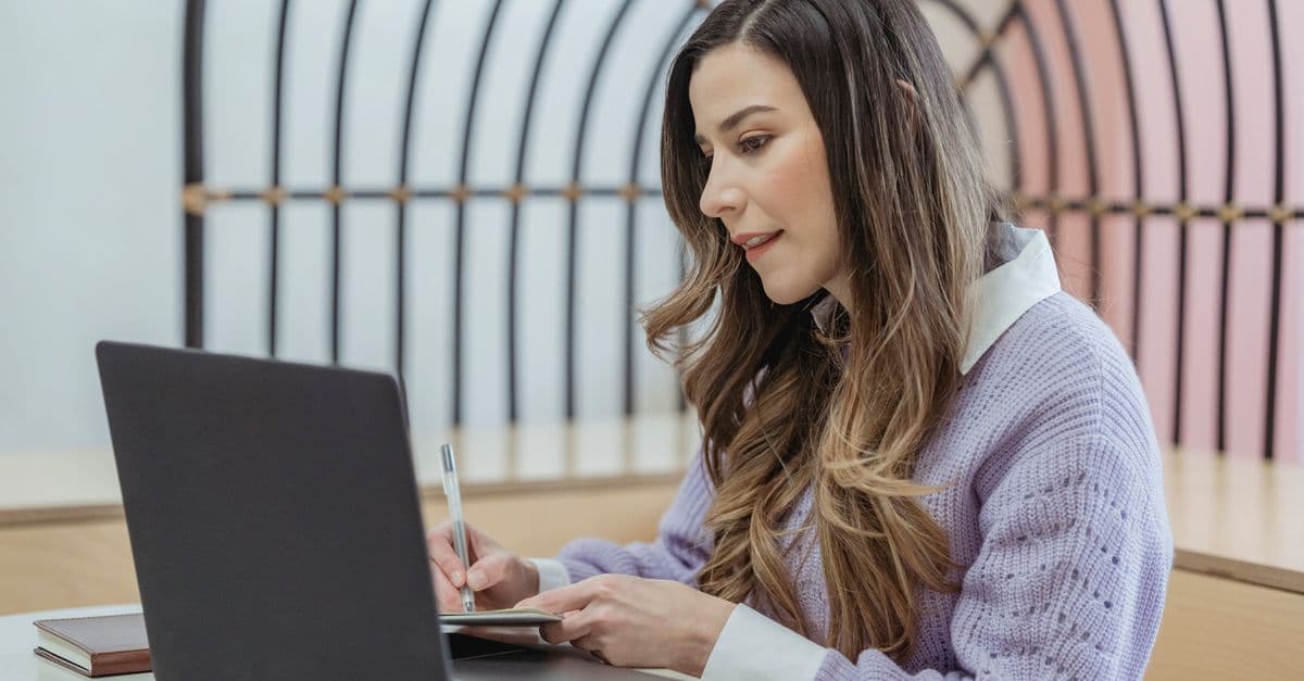 Focused female freelancer with wavy hair writing notes in planner while sitting at table and working on netbook