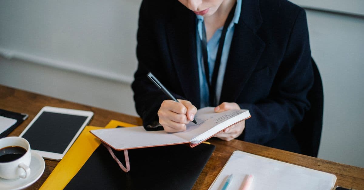 High angle of crop female in formal wear sitting at table and writing notes in planner during work day
