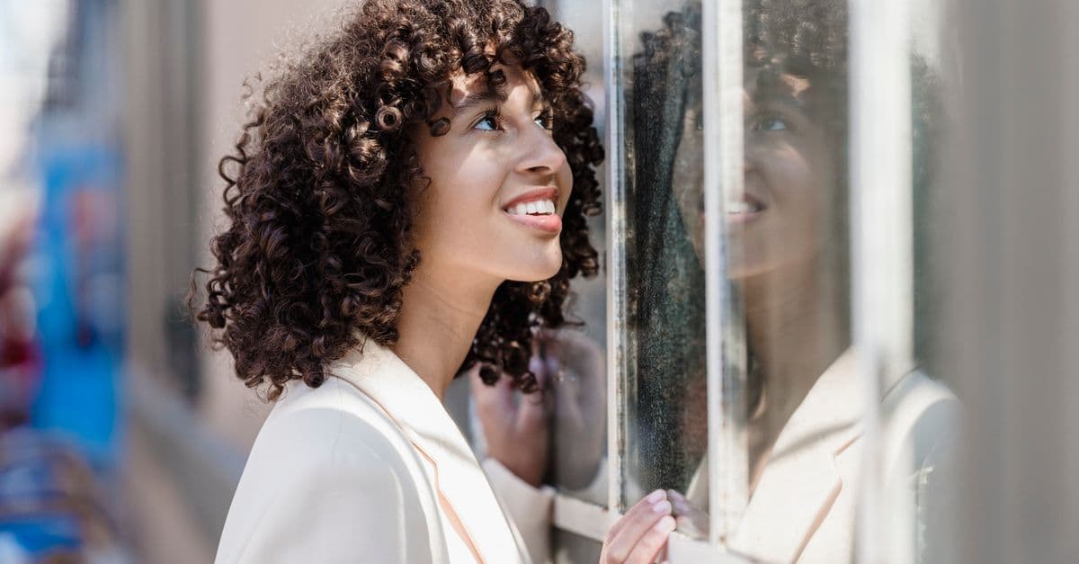 Side view of positive ethnic female with curly hair standing near window and looking up with interest