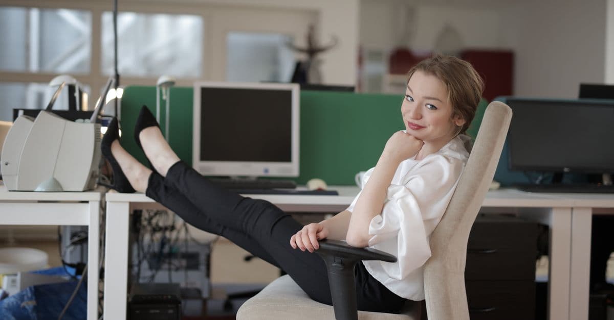 Side view of cheerful female worker in formal style relaxing on office chair during break while sitting with feet on table and looking at camera