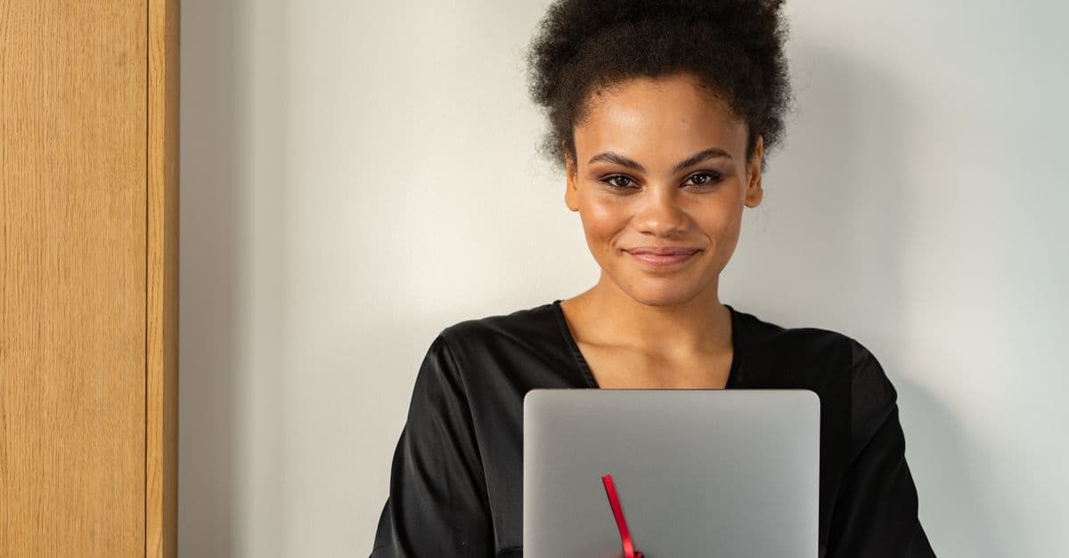 Confident woman indoors holding a laptop and smiling warmly.