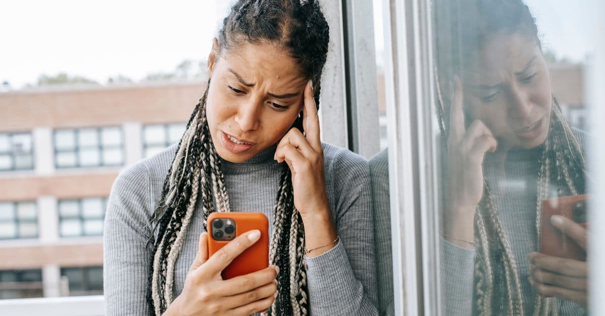 Unhappy African American female standing on balcony while holding smartphone and looking at screen