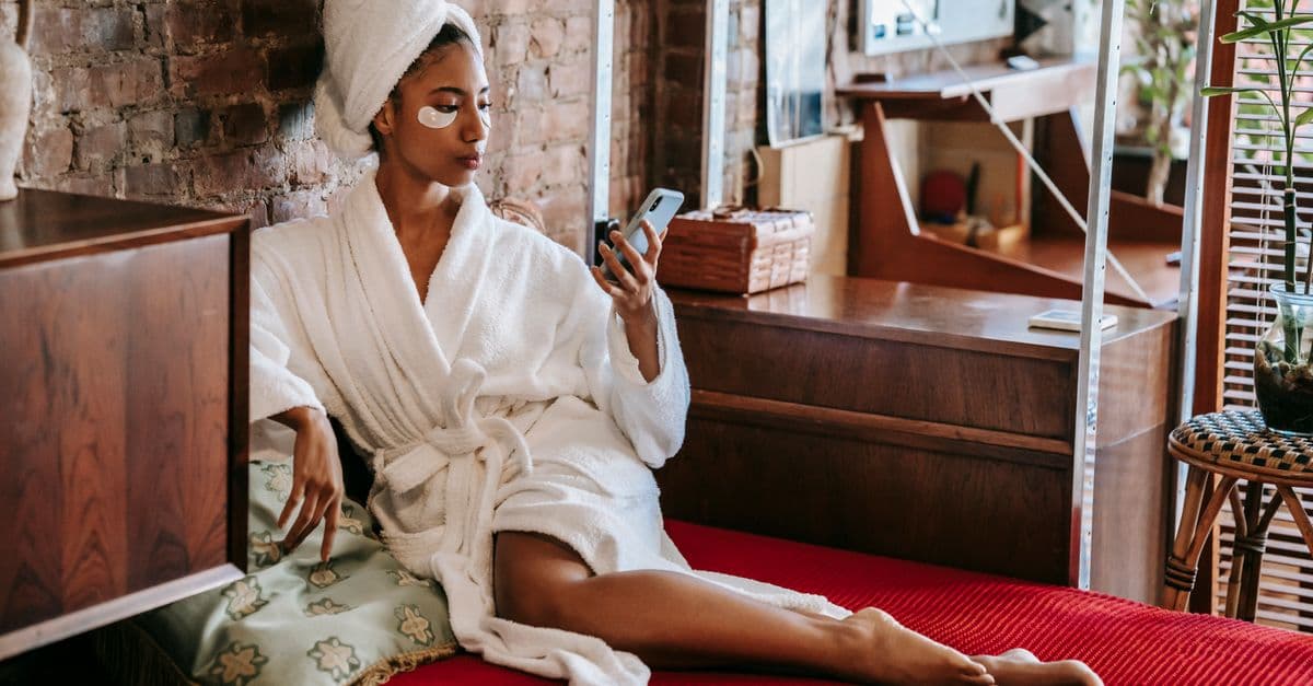 Young African American female in bathrobe sitting on bed and browsing smartphone while resting in cozy room